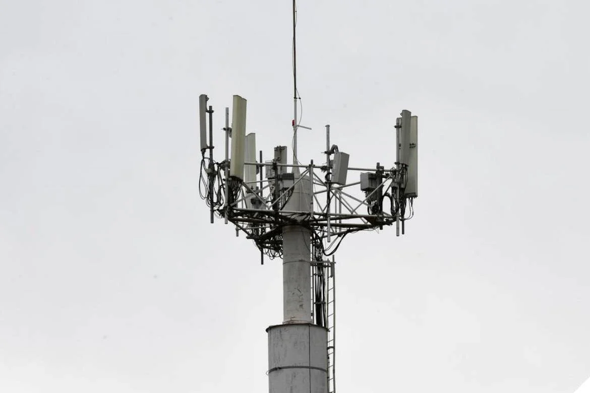 rede 5G no Brasil: Torre de telefonia celular com antenas setoriais e equipamentos no topo, contra céu cinza claro