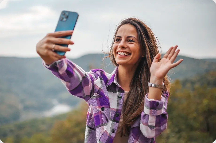 Mulher de camisa xadrez roxa sorri e acena para o celular, numa chamada de vídeo ou selfie, ao ar livre diante de montanhas.